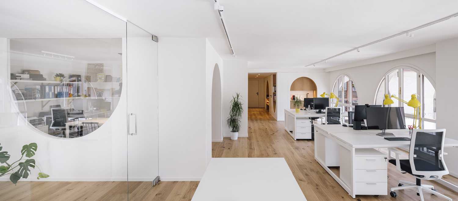 Zooco Studio présente son bureau en espace ouvert où les fenêtres en arc sont le point de départ pour concevoir l'espace à travers des meubles en bois. La forme courbée est mise en évidence par le bois tandis que le blanc définit les espaces minimalistes, un contraste qui génère du mouvement